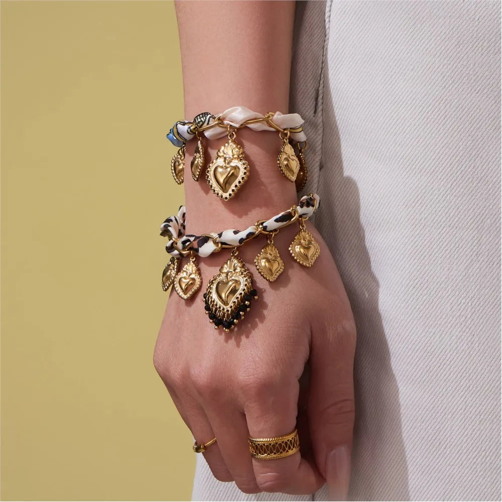 Hand wearing multiple gold bracelets with heart-shaped charms against a neutral background